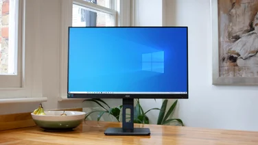 The AOC Q27P2CA 27in monitor on a table with a white wall, painting, plant and windows in the background