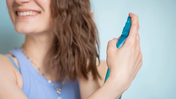 A smiling woman sprays her hair with a Sea Salt spray