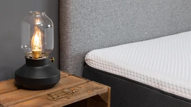 The corner of a hybrid mattress next to a bedside table with a lamo
