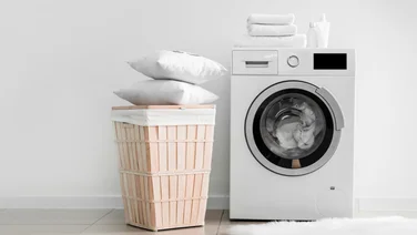 washing machine with clothes on top and a wicker basket beside it with pillows on top, on a white domestic background