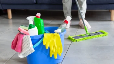 Cleaning tips - featured. Bucket with gloves, cloth and cleaning supplies in foreground with the legs of someone mopping in the background