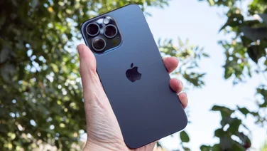 iPhone 15 Pro Max in hand, rear view, in front of some trees