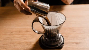 A person pours ground coffee over a clever coffee dripper on a wooden work surface