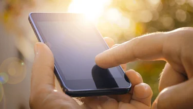 How to prevent your phone overheating in the sun - featured. Close up of hands holding phone, with one finger tapping the screen. The sun is shining brightly in the background