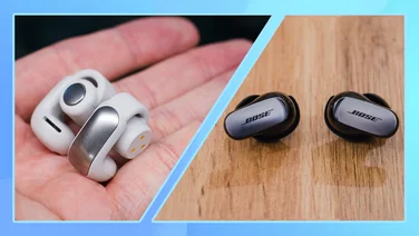An image showing a hand holding the Bose Ultra Open Earbuds on the left hand side, and the Bose QC Ultra Earbuds displayed on a wooden surface on the right