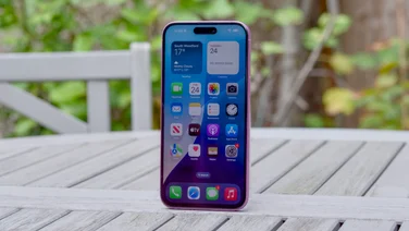 The front of an Apple iPhone 16 Plus on a grey table outside with plants in the background