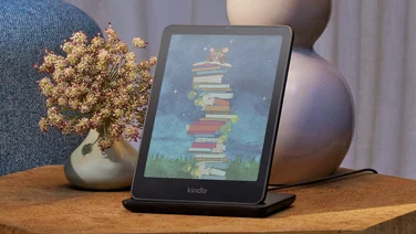 Amazon Kindle Colorsoft sitting on the charging dock on a bedside table, showing a colourful stack of books on the display