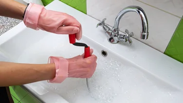 Person wearing pink rubber gloves uses drain snake on blocked sink