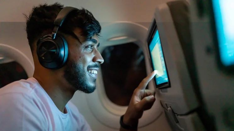 A man wearing wireless headphones using the in-flight entertainment system on the back of an aeroplane seat