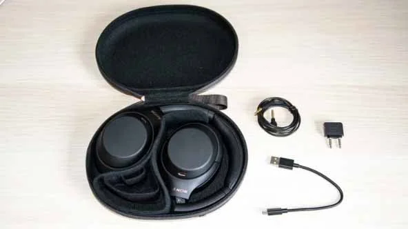 The Sony WH-1000XM4 over-ear headphones in their carrying case with accessories alongside, including a two-pin aeroplane adapter