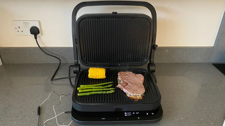 The Zwilling Enfinigy Contact Grill, open, on a kitchen countertop, with steak, corn and asparagus cooking on its surface