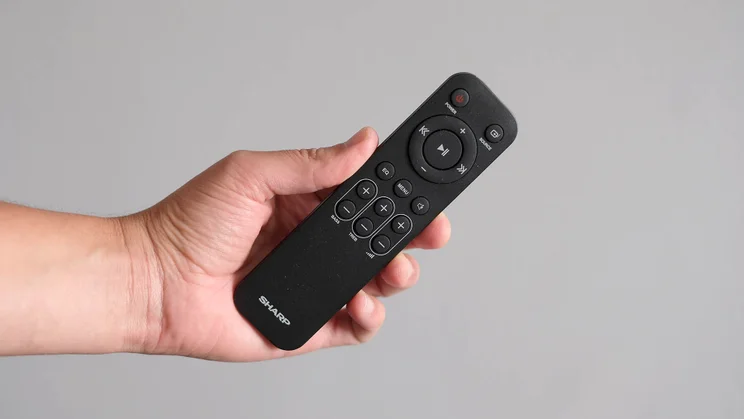 The remote control included with the Sharp HT-SBW55121 soundbar being held in a man's hand against a grey wall