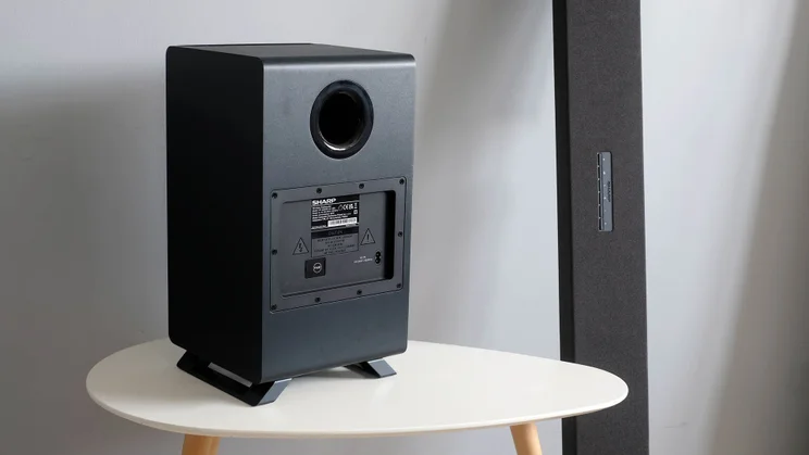 Rear view of the subwoofer component of the Sharp HT-SBW55121 sound system on a wooden table, with the soundbar leaning against the wall next to it
