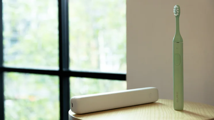 The Suri 2.0 electric toothbrush next to its charging case, on a stool by a window