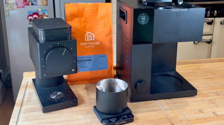 The Fellow Aiden coffee machine pictured on a kitchen table next to a Fellow coffee grinder and coffee mug