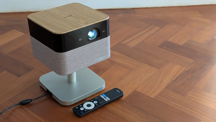 The Epson Lifestudio Flex EF-72 4K projector on a wooden floor with its remote control next to the in-built stand