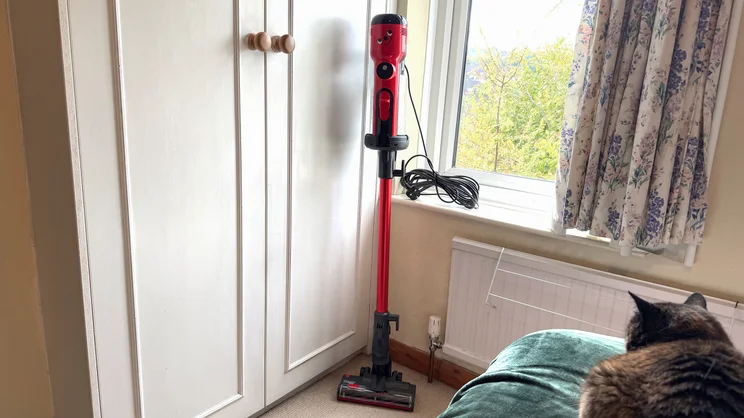 The Henry Quick Corded vacuum cleaner stood up in the corner of a room by a window. A cat looks on from a bed.