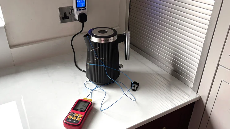 The Russell Hobbs Bronte kettle on a kitchen worktop. A thermometer and temperature probes are being used to monitor internal and external temperature. The kettle is plugged into a mains socket via a plug-through power meter