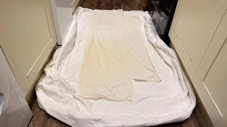 The Dusk Luxury Quilted Mattress Protector on a floor, with a piece of bedsheet over the top