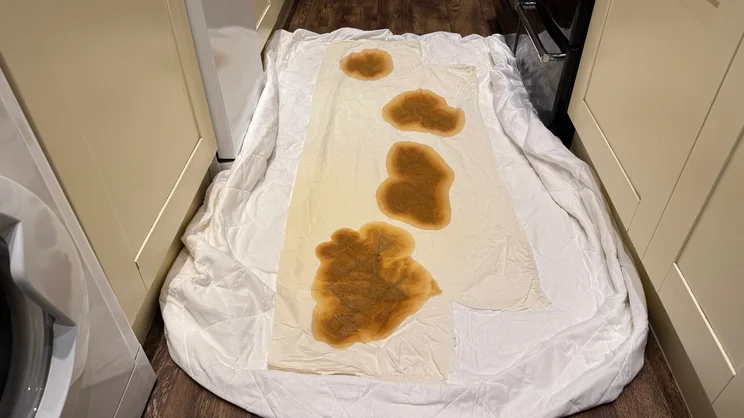 The Dusk Luxury Quilted Mattress Protector on a floor, with a piece of bedsheet over the top. Several coffee stains are covering the sheet