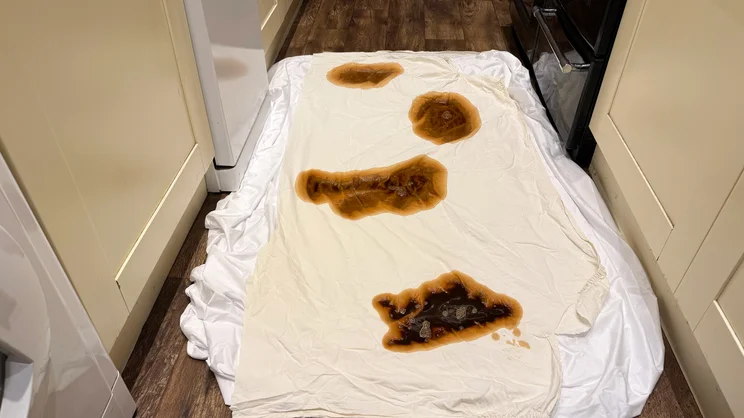 The Nectar mattress protector on the floor, with a polycotton sheet covering it. Several coffee stains and small puddles are covering the sheet