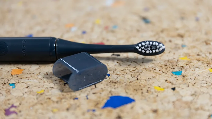 The Ordo Sonic Edge toothbrush laid down on a multicoloured cork table, alongside travel cap