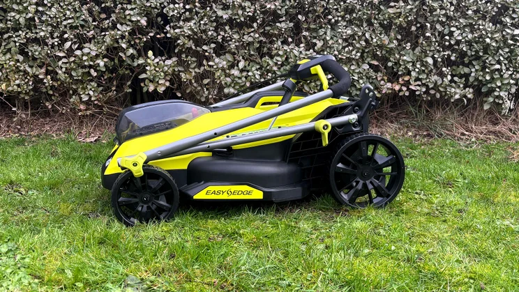 The Ryobi RY18LMX33A lawn mower folded, on a lawn in front of a hedge