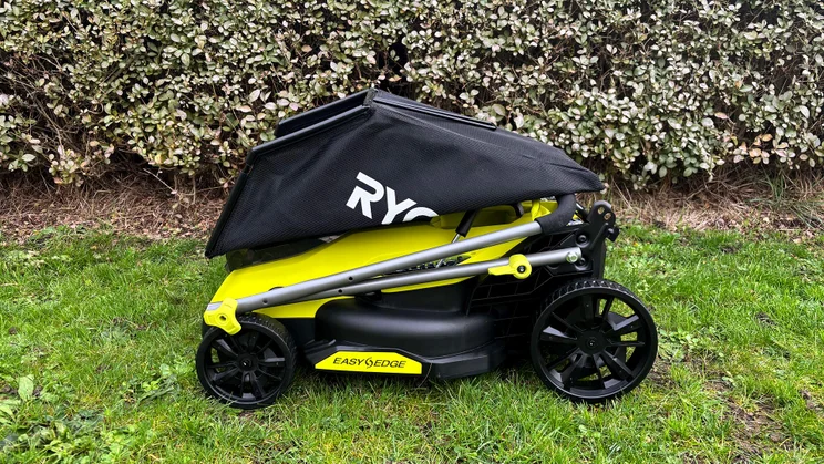 The Ryobi RY18LMX33A lawn mower folded, with grass collector on top, on a lawn in front of a hedge