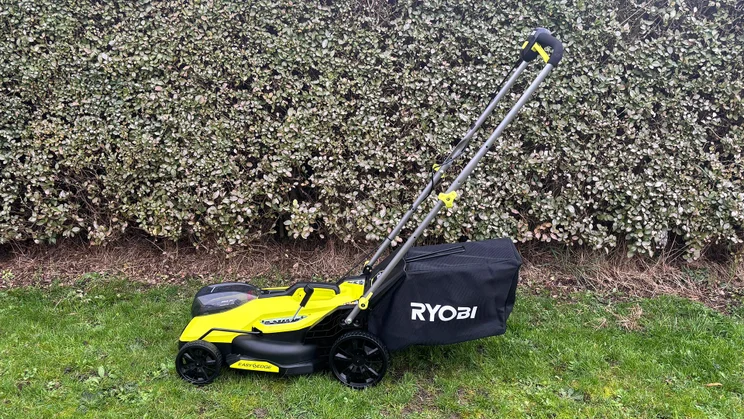 A side view of the Ryobi RY18LMX33A lawn mower, on a lawn in front of a hedge