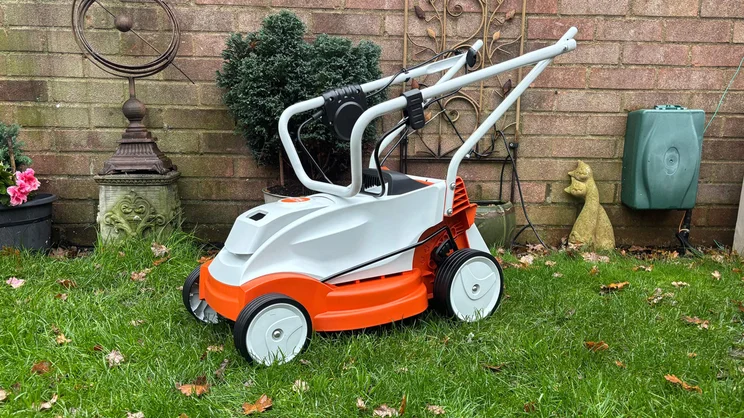 The Stihl RMA 235 lawn mower, with its handles folded, in a garden in front of a brick wall