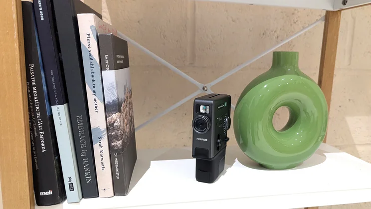 The Instax Mini Evo Cinema on a white shelf, between books and a green vase