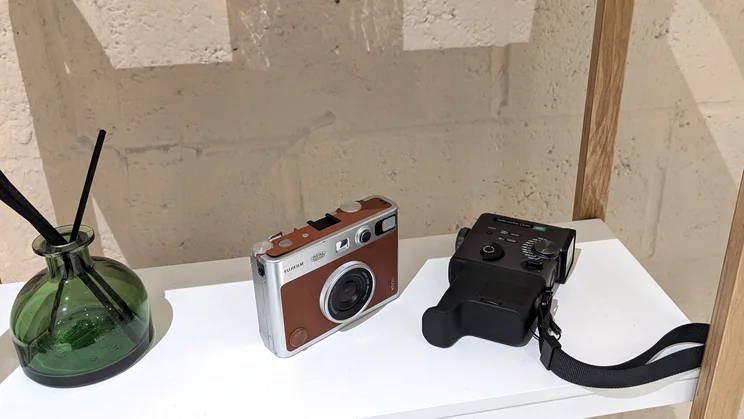 The Instax Mini Evo Cinema camera on a white shelf, next to the Instax Mini Evo camera and a green reed diffuser