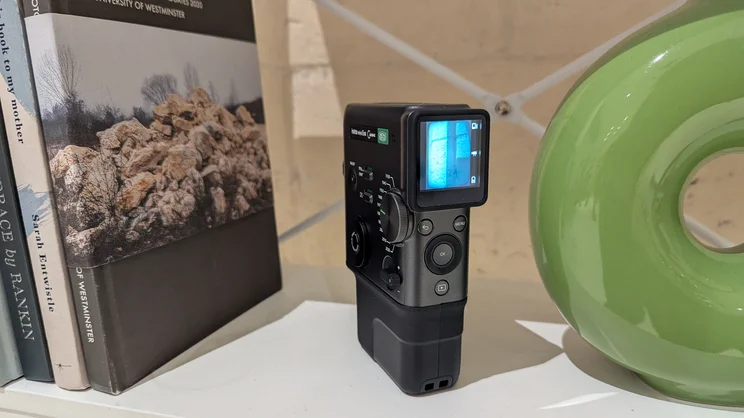 The Instax Mini Evo Cinema on a white shelf, between books and a green vase