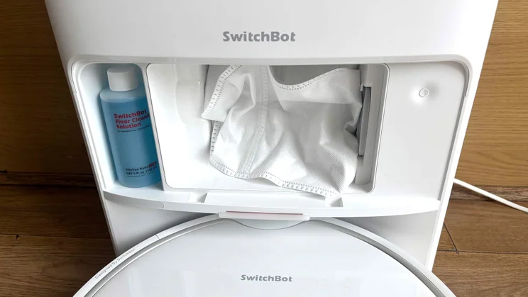 The Switchbot S20 robot vacuum's dock with front panel removed, showing the dust bag and a bottle of cleaning solution