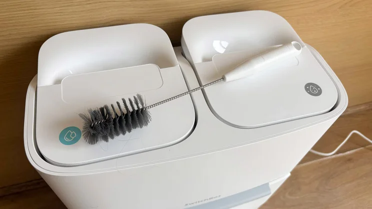 The top of the Switchbot S20 robot vacuum's dock and base, showing water tanks. The vacuum's detangling tool is placed on top