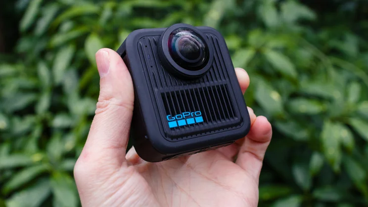 The GoPro Max 2 held at an angle, pictured from the front with greenery in the background