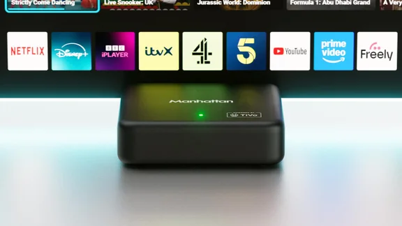 The Manhattan Aero 4K TV Streamer in front of a TV displaying a row of apps, including Freely