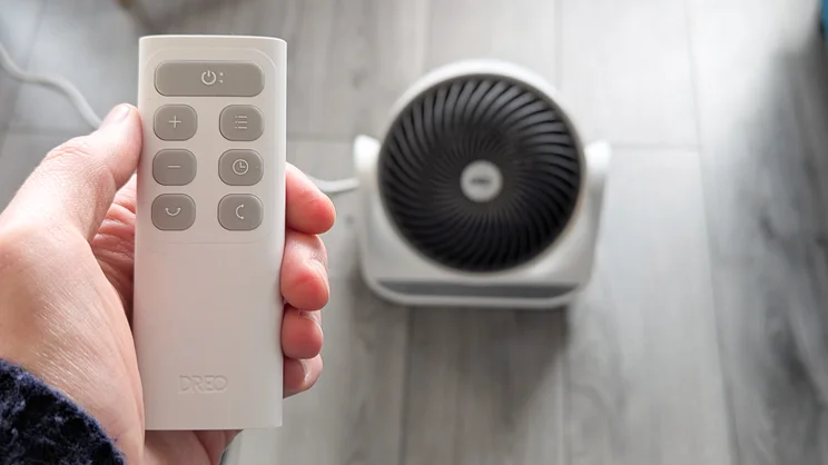 A hand holding the Dreo Dreo Smart Whole Room Heater 714s remote control. The heater is on the floor in the background