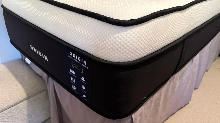A closeup of a corner of the Origin LumbarCloud mattress, on a bedframe. The company's logo is on the bottom side of the mattress