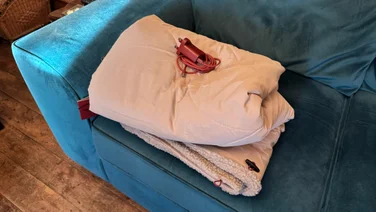 The Stoov Huge Hug 3 electric blanket folded up on a blue sofa. Its USB-C adapter is placed on top of it