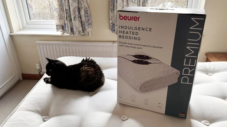 The Beurer Indulgence Heated Underblanket in its box, on top of a mattrress, next to a cat