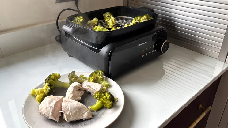 A plate of steamed chicken and broccoli, on a kitchen surface, in front of the Panasonic 6-in-1 Table-top Multi Cooker