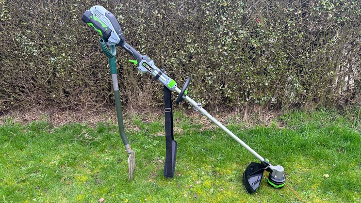 The Ego ST1511E-T grass trimmer propped upright with a spade, on a lawn in front of a hedge