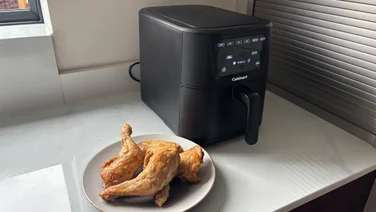 A plate of cooked chicken legs in front of the Cuisinart Compact Max Air Fryer, on a kitchen worktop
