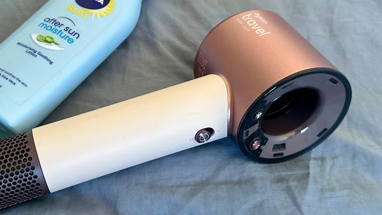A closeup of the Dyson Supersonic Travel hair dryer, lying on a blue bedsheet alongside a bottle of aftersun