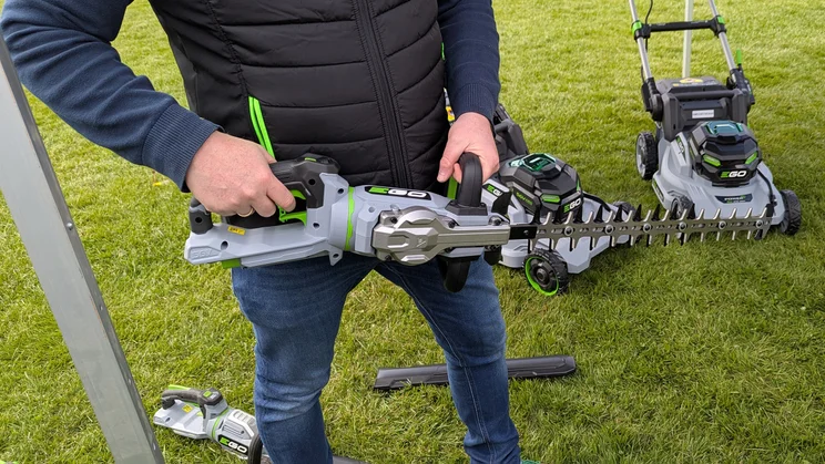 An EGO hedge trimmer is held during a demonstration. In the background are two EGO lawn mowers