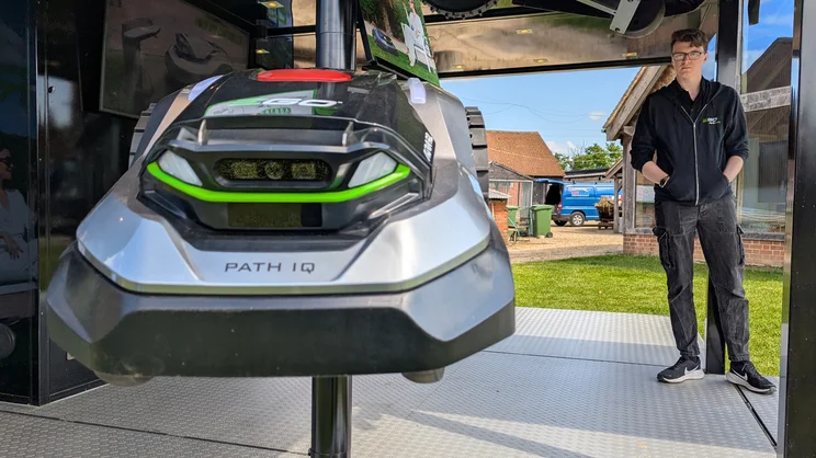 A closeup in the EGO Aura R2 robot lawn mower on a display stand. In the back right, a man looks on at it