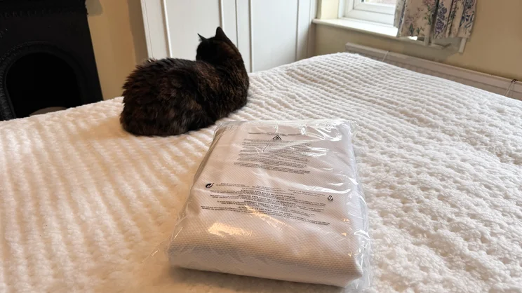 The Emma Mattress Protector in its plastic packaging, on a mattress. A cat is sitting next to it, looking away from the camera