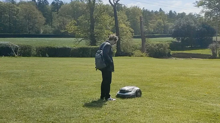 Expert Reviews' Home Editor Gareth Thomas stands in front of an EGO Aura R2 robot lawn mower, on a farm lawn