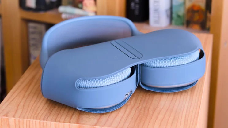 The Apple AirPods Max 2 in their case on top of a speaker with a bookshelf in the background